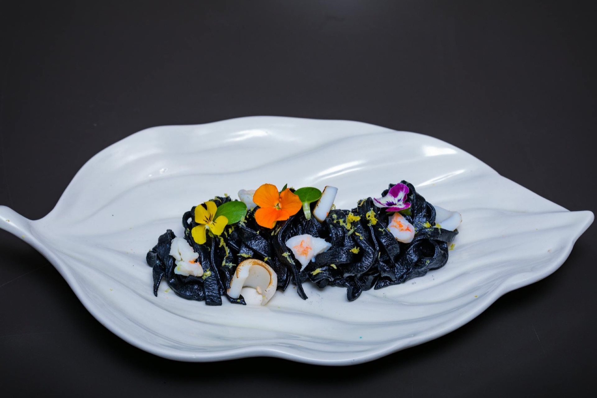 Pâtes noires à l'encre de seiche avec fleurs comestibles violettes et orange sur plat blanc en forme de feuille