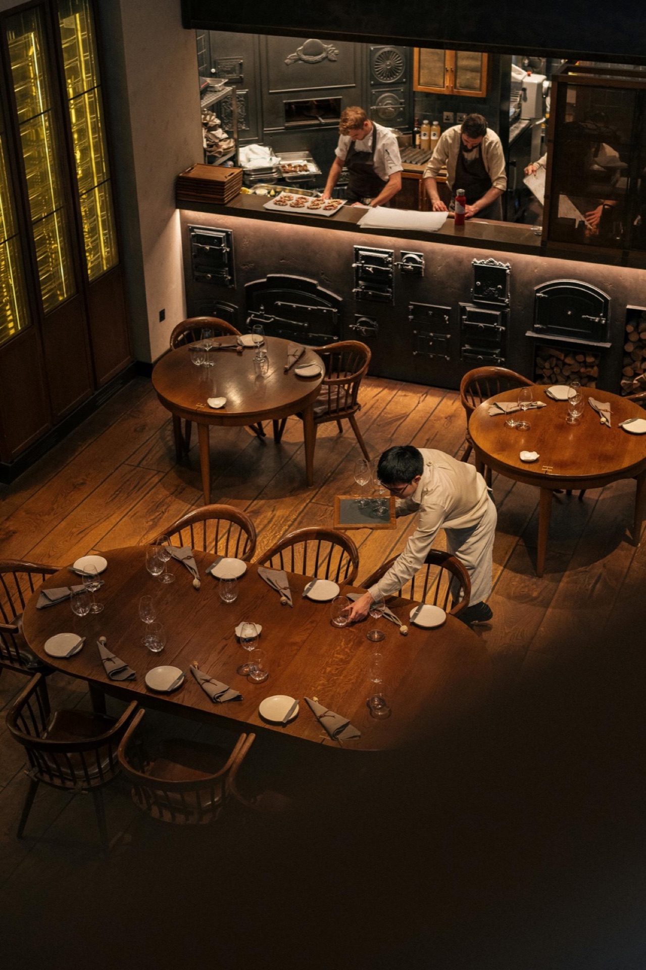 Salle de restaurant intime, cuisine ouverte avec chefs, tables en bois et chaises arrondies
